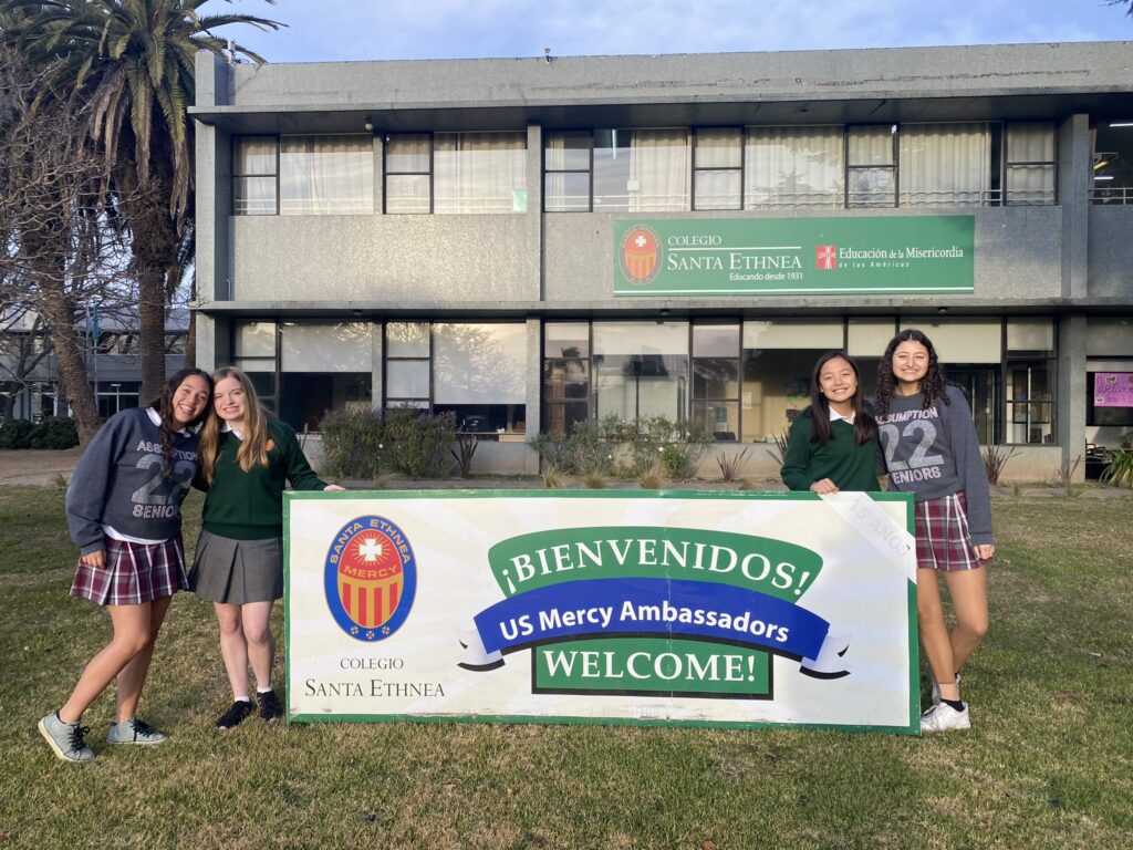 The authors of this article standing next to a banner welcoming them to Colegio Santa Ethnea as Mercy Ambassadors